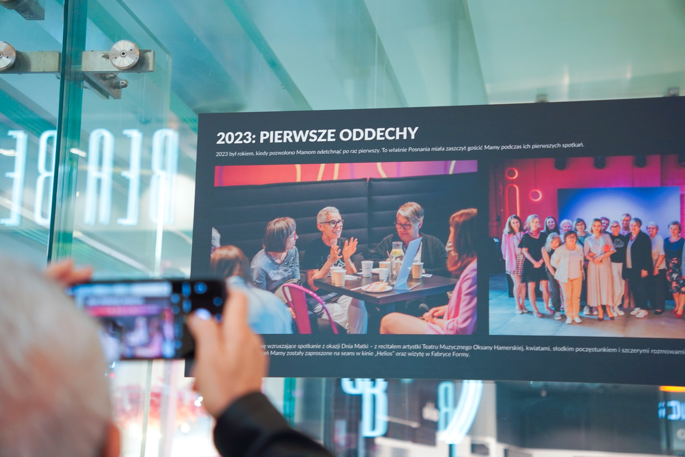 Kobieta robi zdjęcie planszy wystawy prezentującej fotografie z projektu Oddech dla Mam. Obok niej stoją inne uczestniczki wydarzenia.